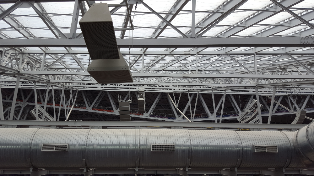 This image captures the intricate interior roof structure of US Bank Stadium in Minneapolis, MN. The roof is composed of a complex web of white steel trusses supporting a translucent roof that allows natural light to filter through. A sizable cylindrical air duct runs horizontally across the image, featuring vents and reinforcing bands.