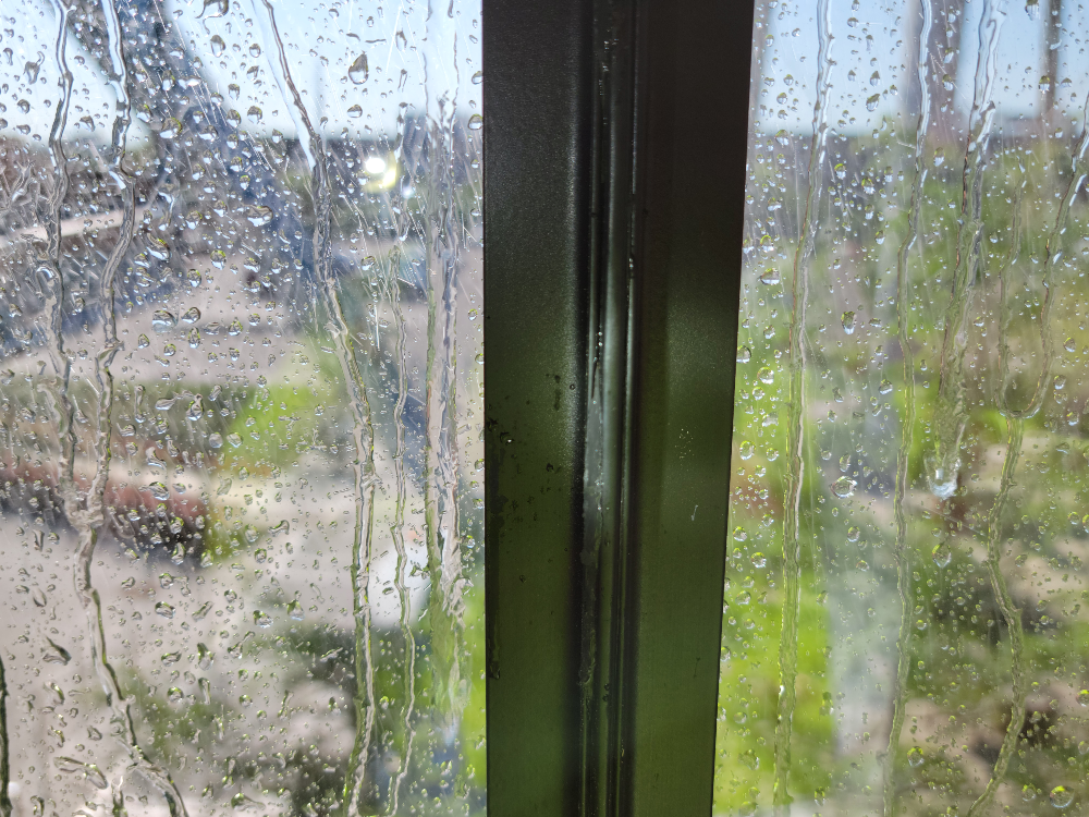 This image features a close-up view of a windowpane with water condensation. Droplets of water are scattered across the glass, and streaks of moisture run vertically down the pane, creating a textured effect that slightly distorts the view.