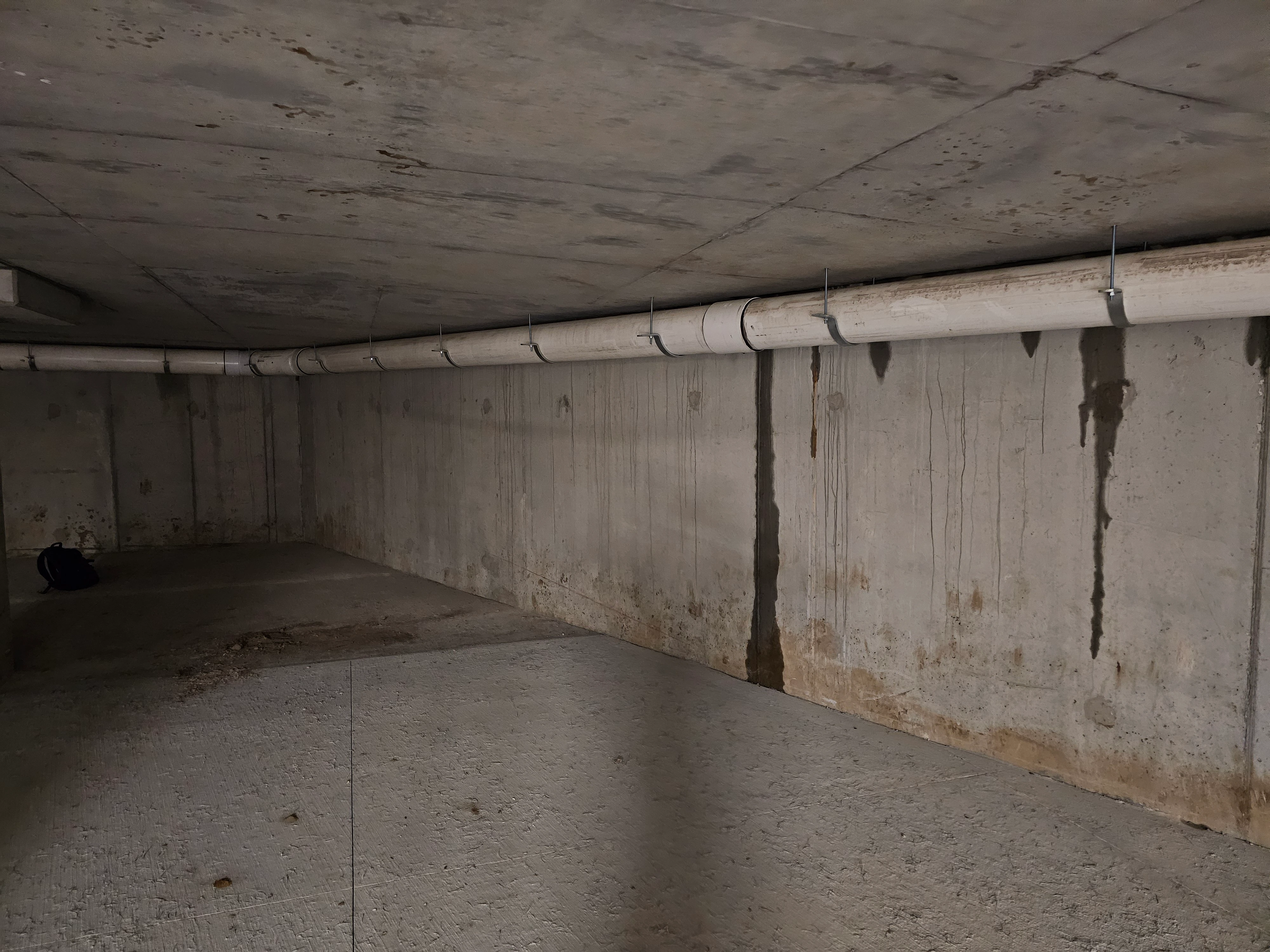 A concrete space with visible water stains and possible signs of moisture damage on the walls and floor. A large white PVC pipe runs horizontally along the upper portion of the wall, supported by metal brackets. The concrete surfaces appear damp, with discoloration and streaks where water has likely dripped or seeped over time.