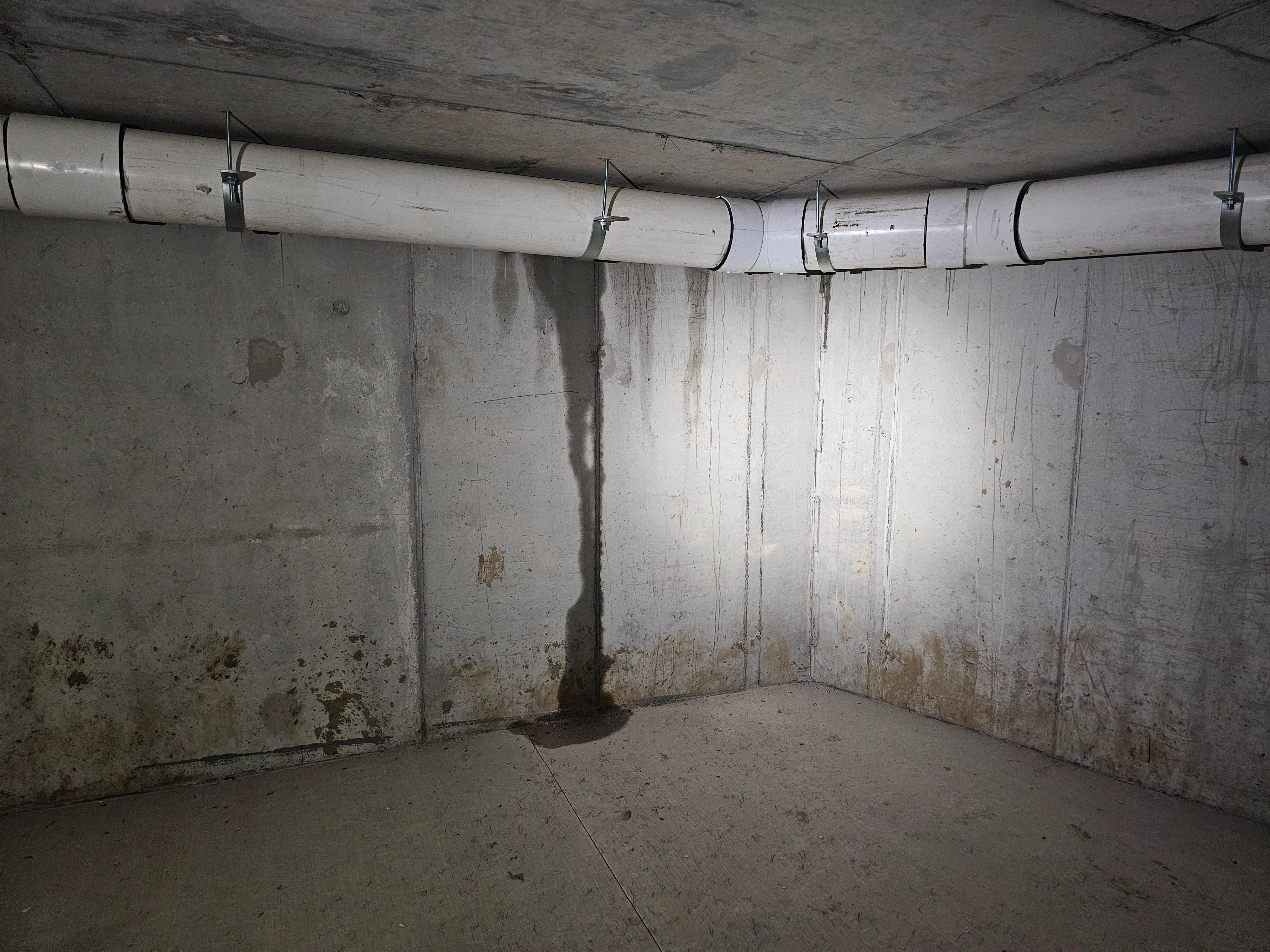 A basement or a crawl space with visible water stains and possible signs of moisture damage on the walls and floor. A large white PVC pipe runs horizontally along the upper portion of the wall, supported by metal brackets. The concrete surfaces appear damp, with discoloration and streaks where water has likely dripped or seeped over time.