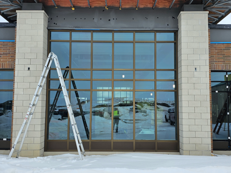 This image depicts a large glass curtain wall framed by light beige brick columns on a building's exterior. Above the glass wall, exposed dark brown beams under construction indicate that the building is undergoing fenestration curtain wall testing.